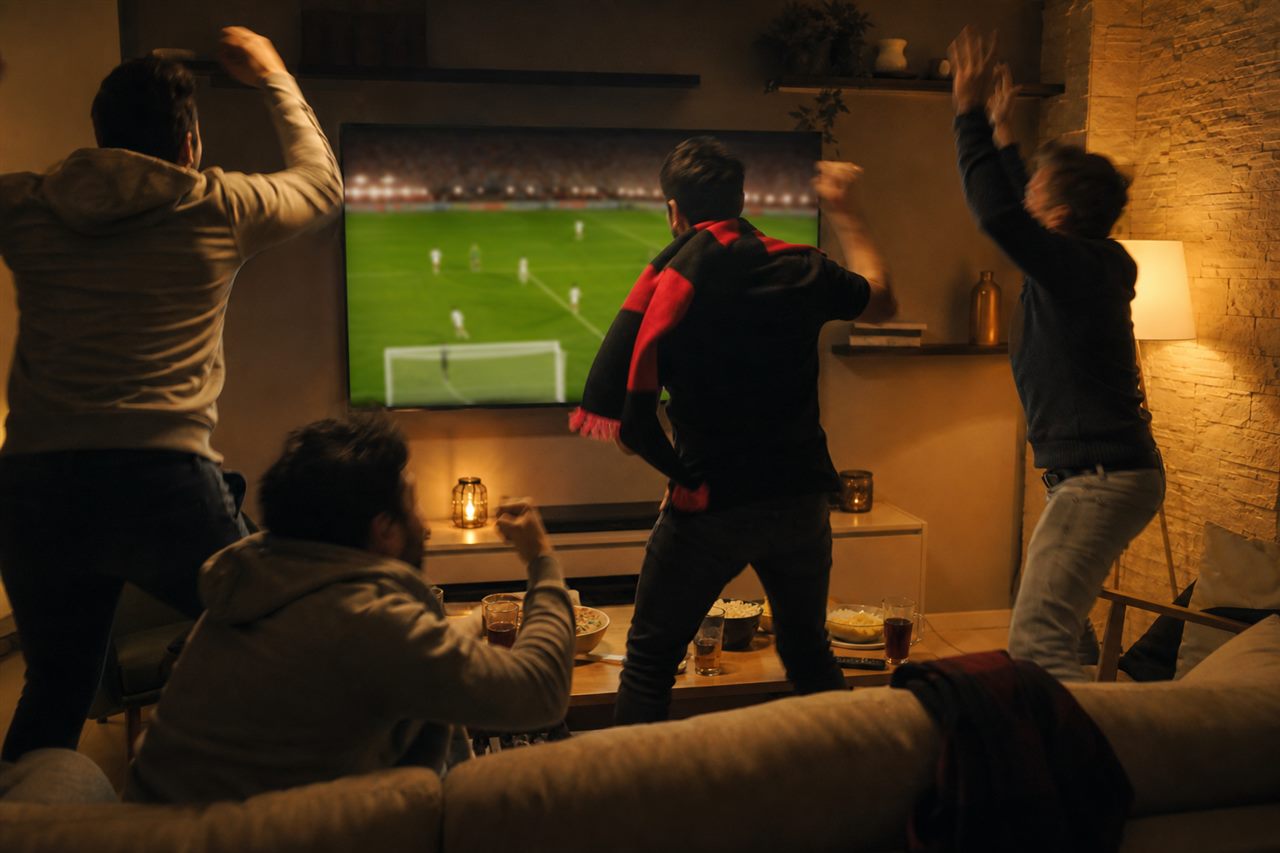 A small group of friends in a cozy living room late at night leaping up in shock and joy as they react to a dramatic late goal on the television, warm lamp light, one draped in a plain black and red scarf, no identifiable faces
