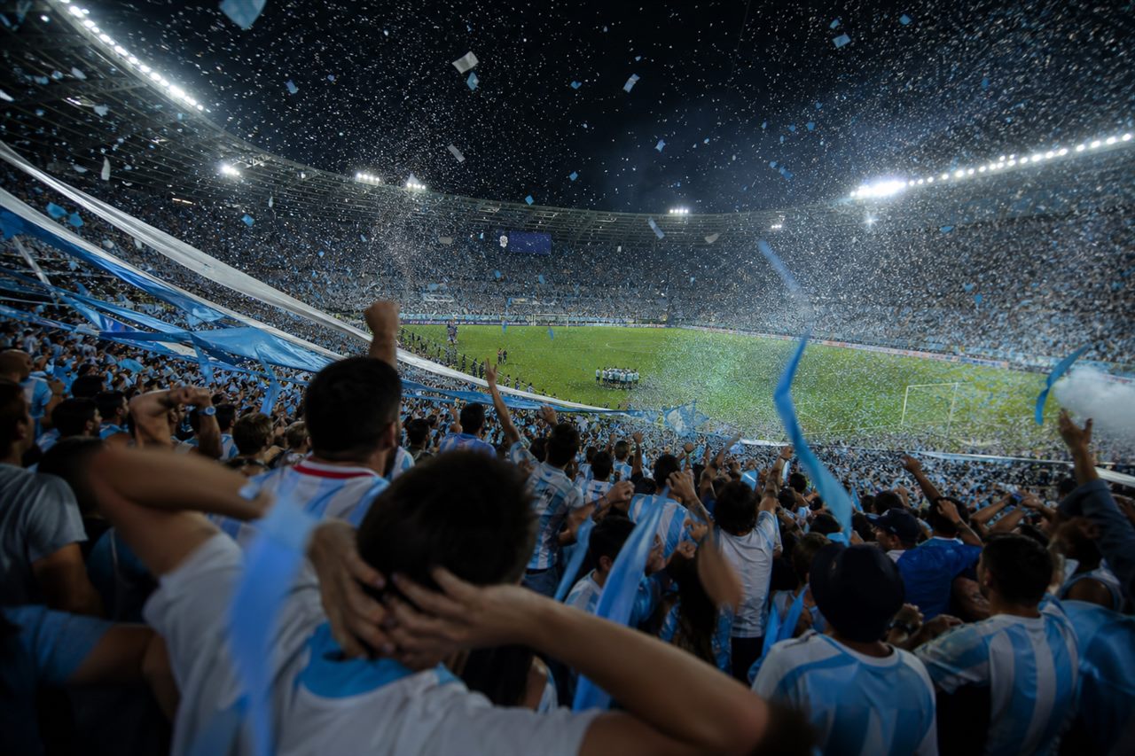 A packed night stadium as supporters in light blue and white erupt in relief and joy at a late comeback winner, arms raised, confetti drifting in the floodlights, no identifiable faces, no readable text or logos