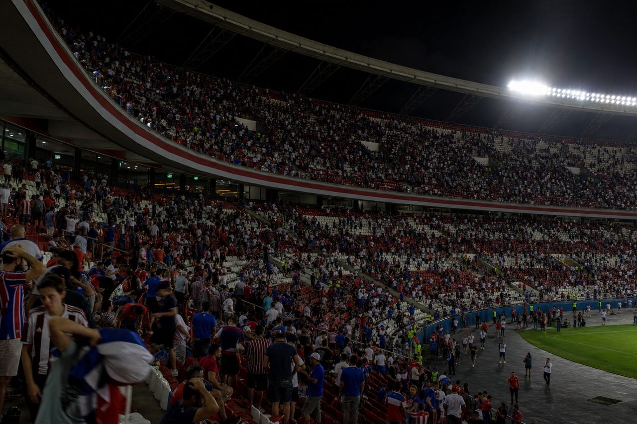 A packed night stadium as home supporters in red white and blue sit dejected with heads in their hands after a heavy defeat, some already leaving their seats, a home tournament dream ending, no identifiable faces, no readable text or logos